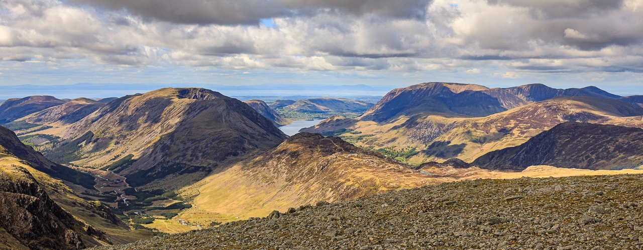 A view across the tops of numerous fells, with the Solway Firth and Scotland in the
far distance. The fell tops are lit up with dappled sunlight as the clouds visible above
create shadows across the landscape
